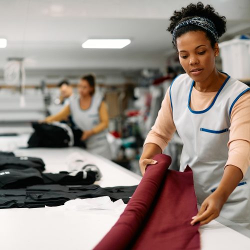 Young African American seamstress working with fabric at clothing design workshop.